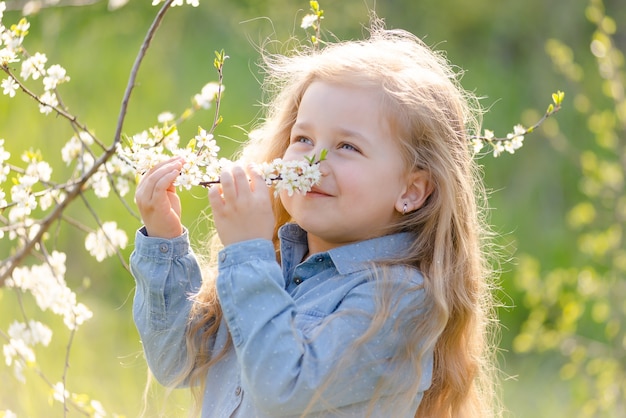 Niña rubia linda con el pelo largo huele una rama de árbol en flor en el parque en primavera.