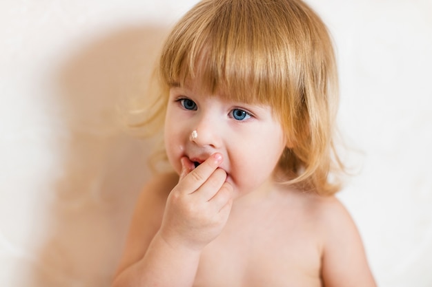 Niña rubia en pantalón rosa sentado en la mesa blanca comiendo pastel de cumpleaños