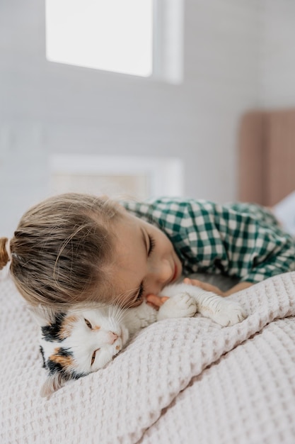 Una niña rubia con pijama abraza a un gato doméstico en su dormitorio el concepto de una acogedora mañana en casa