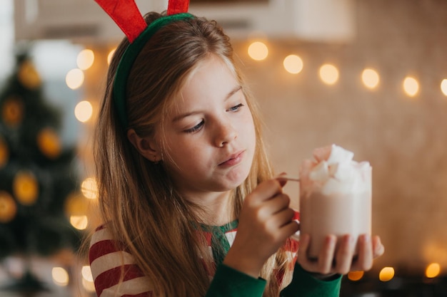 Niña rubia en pijama de rayas sostiene una taza con bebida caliente y malvaviscos en la cocina decorada. fines de semana y festivos de invierno. lugar para el texto.