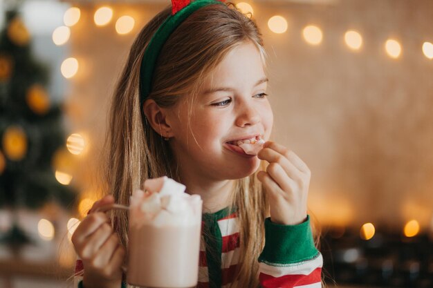 Niña rubia en pijama de rayas sostiene una taza con bebida caliente y malvaviscos en la cocina decorada. fines de semana y festivos de invierno. lugar para el texto.