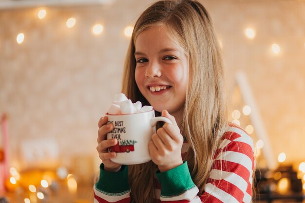 Niña rubia en pijama de rayas sostiene una taza con bebida caliente y malvaviscos en la cocina decorada. fines de semana y festivos de invierno. lugar para el texto.