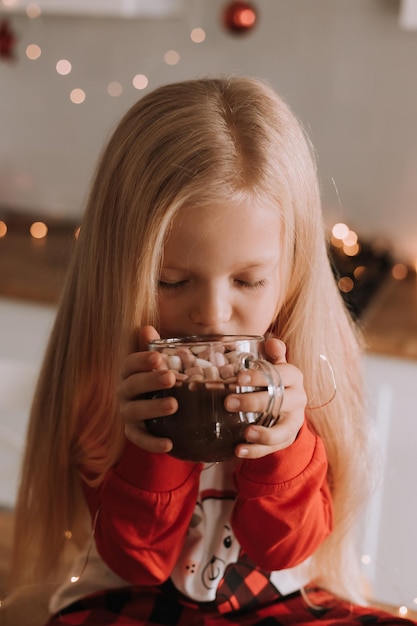 Niña rubia en pijama rojo de Navidad bebe cacao con malvaviscos en una cocina decorada. acogedora casa de invierno los fines de semana y vacaciones. estilo de vida. espacio para texto. foto de alta calidad