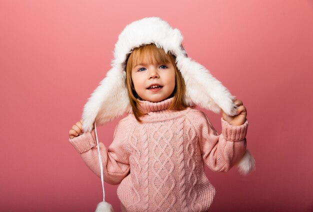 Niña rubia en ropa de invierno y un sombrero de piel sobre un fondo rosa.