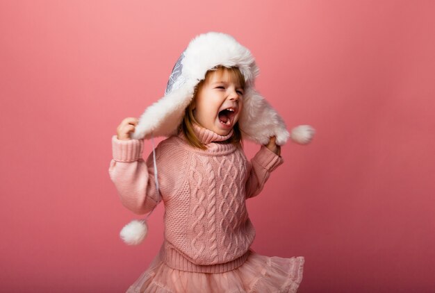 Niña rubia en ropa de invierno y un sombrero de piel sobre un fondo rosa.