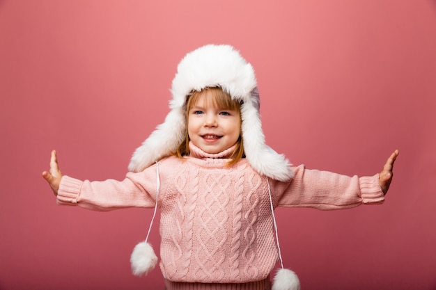 Niña rubia en ropa de invierno y un sombrero de piel sobre un fondo rosa.