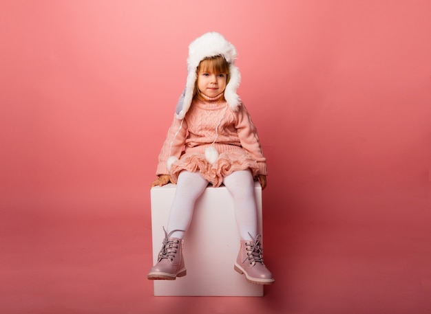 Niña rubia en ropa de invierno y un sombrero de piel sobre un fondo rosa.