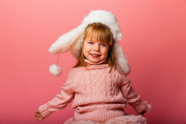 Niña rubia en ropa de invierno y un sombrero de piel sobre un fondo rosa.