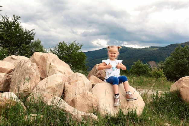 Niña rubia sentada en una piedra grande sobre un fondo de montañas y muestra los dedos hacia arriba