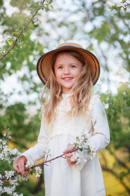 Niña rubia en un sombrero de paja cerca de un árbol floreciente