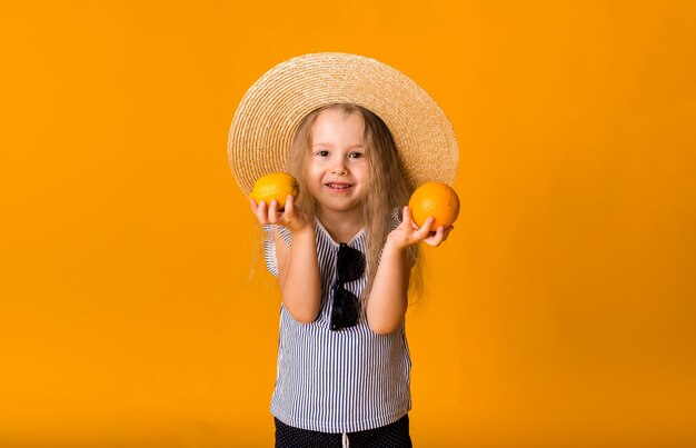 Una niña rubia con un sombrero de paja sostiene una naranja y un limón sobre una superficie amarilla con espacio para texto