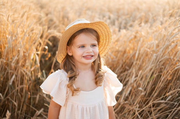 niña rubia con sombrero de paja y vestido de muselina beige en campo de trigo al atardecer