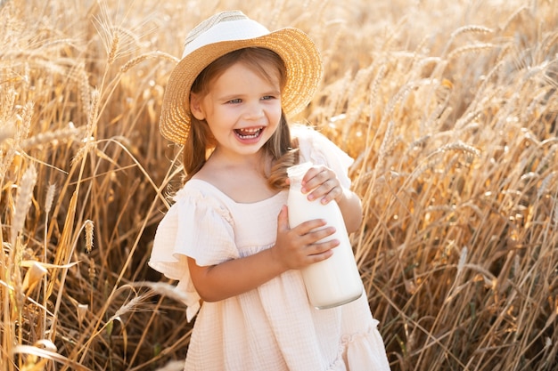 Niña rubia con sombrero de paja y vestido de muselina beige sostiene la batalla de la leche en el campo de trigo al atardecer