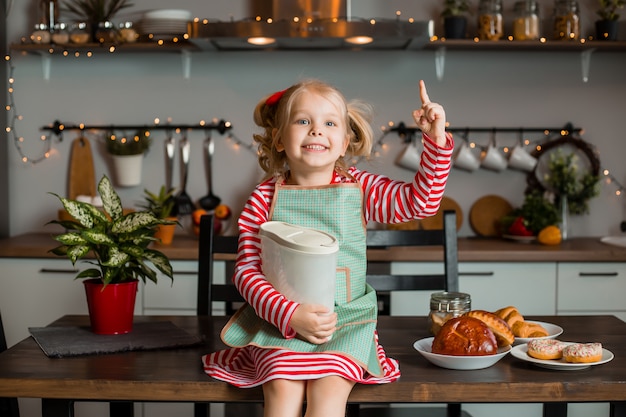 Niña rubia sonríe en la cocina, cocina un pastel. Niño en la cocina. Quédate en casa