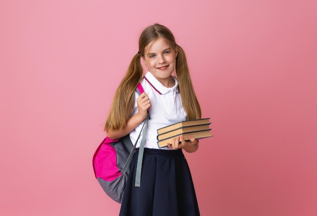 Niña rubia sonriente de 1012 años en uniforme escolar con mochila sosteniendo libros aislados en un retrato de estudio de fondo rosa pastel El concepto de estilo de vida infantil Educación en la escuela