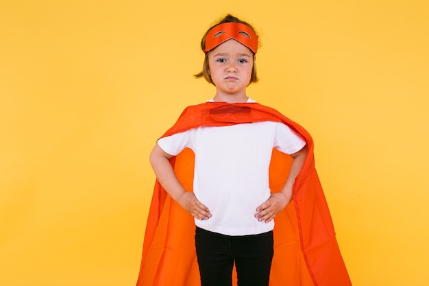 Niña rubia vestida como un superhéroe superheroína con una capa y una máscara roja, enojada, con los brazos en jarras en serio, sobre fondo amarillo