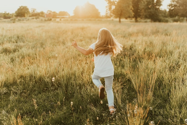 una niña rubia vestida con ropa de colores claros y una camiseta blanca se escapa de la burbuja de jabón