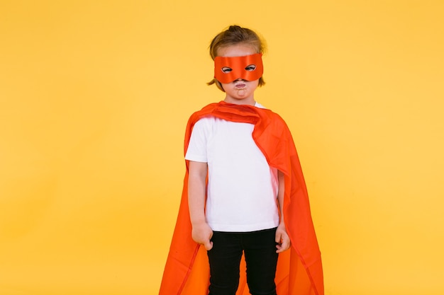 Niña rubia vestida de superhéroe superheroína con capa y máscara roja, enojada, con la capa al lado, sobre fondo amarillo