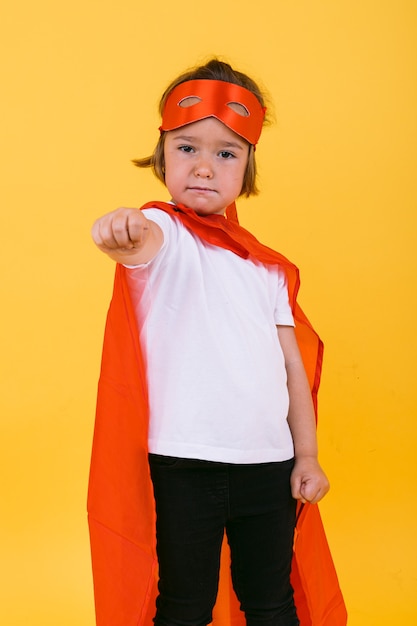 Foto niña rubia vestida de superhéroe superheroína con capa y máscara roja, levantando su brazo en posición de vuelo, sobre fondo amarillo