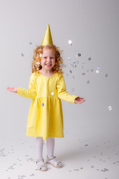 Niña rubia en un vestido amarillo atrapa confeti sonriendo feliz en el espacio en blanco