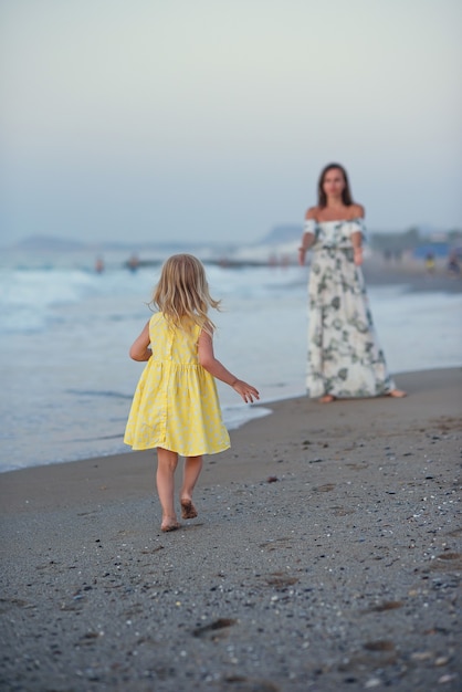 Niña rubia con vestido amarillo corriendo hacia su hermosa madre en la orilla del mar