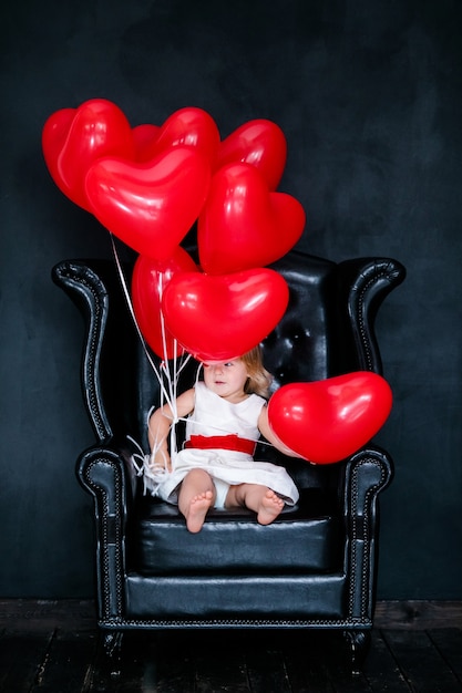 Niña rubia con vestido blanco y cinta roja sentada en el sillón con globo de corazón rojo en el día de San Valentín