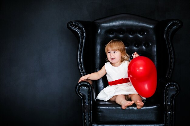 Niña rubia con vestido blanco y cinta roja sentada en el sillón con globo de corazón rojo en el día de San Valentín