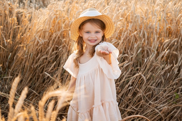 Niña rubia con vestido de muselina beige tiene granos de trigo en la palma de su mano en el campo de trigo al atardecer.