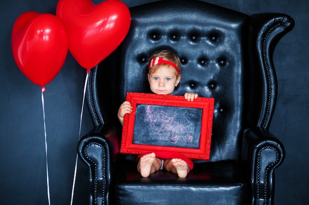 Niña rubia en vestido rojo con corona roja con corazones sentado en el sillón con globo de corazón rojo en el día de san valentín.