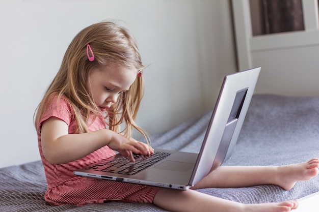 Una niña rubia con un vestido rojo está sentada en la cama con una computadora portátil. Comunicación en línea, videocomunicación, educación.