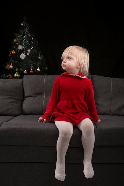 Niña rubia con un vestido rojo se sienta en el sofá con el telón de fondo de un árbol de Navidad y una pared negra y mira hacia un lado. Nochebuena. Año nuevo.