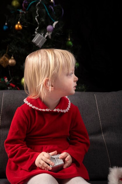 Niña rubia con un vestido rojo se sienta en el sofá con el telón de fondo de un árbol de Navidad y una pared negra y mira hacia un lado. Nochebuena. Año nuevo