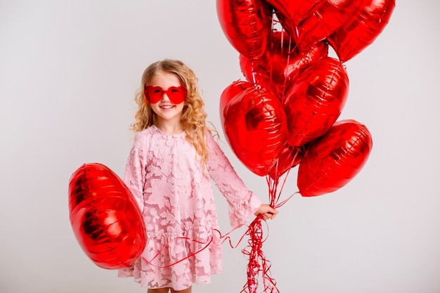 Niña rubia con un vestido rosa está sonriendo y sosteniendo un montón de globos rojos en forma de corazón concepto de día de San Valentín