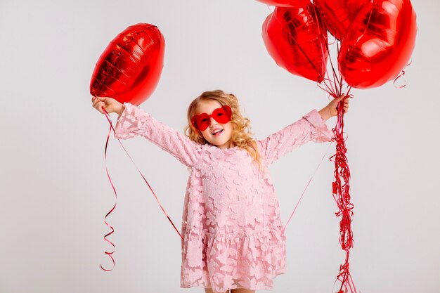 Niña rubia con un vestido rosa está sonriendo y sosteniendo un montón de globos rojos en forma de corazón concepto de día de San Valentín