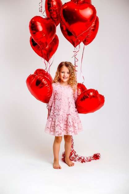 Niña rubia con un vestido rosa está sonriendo y sosteniendo un montón de globos rojos en forma de corazón concepto de día de san valentín