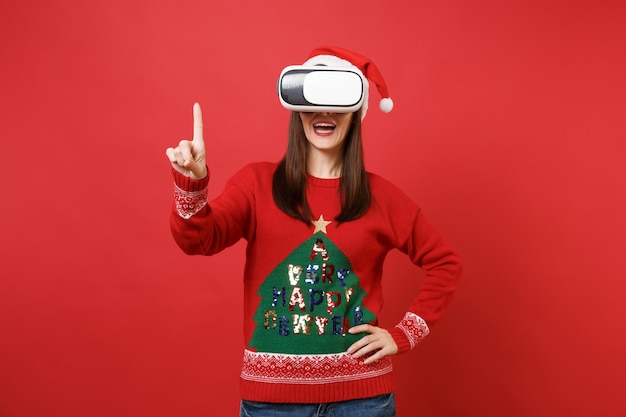 Niña Santa en suéter de punto, sombrero de Navidad, auriculares de realidad virtual, toque algo como presionar, hacer clic en el botón, apuntando a la pantalla virtual flotante aislada sobre fondo rojo brillante de la pared. Decir ah