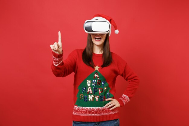 Niña Santa en suéter de punto, sombrero de Navidad, auriculares de realidad virtual, toque algo como presionar, hacer clic en el botón, apuntando a la pantalla virtual flotante aislada sobre fondo rojo brillante de la pared. Decir ah