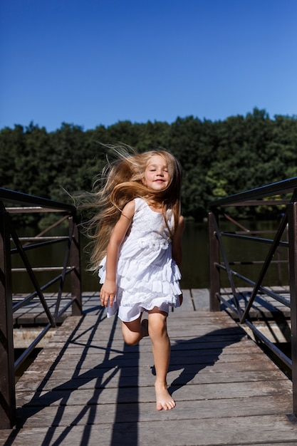 Una niña de seis años lleva un vestido blanco. Hermoso cabello largo. Camina cerca del río en el fondo del bosque.