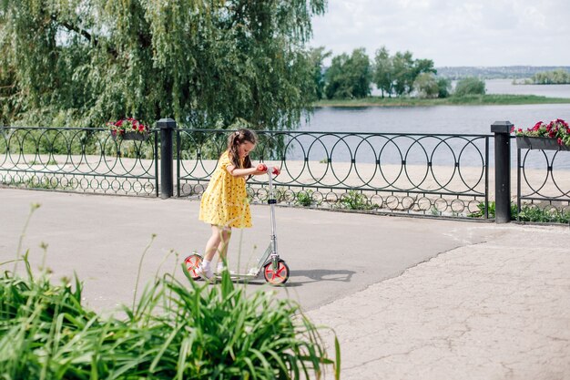 Una niña de seis años con un vestido amarillo está aprendiendo a andar en scooter.