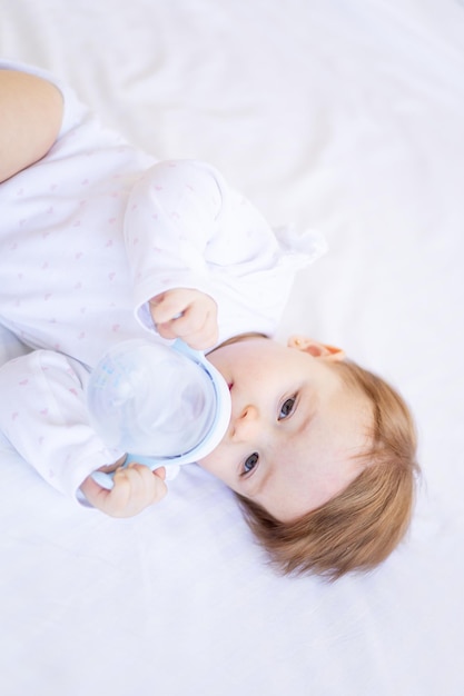 Una niña de seis meses en una cama con ropa blanca chupa una botella de agua o leche. Un niño pequeño en una cama de algodón en casa se despertó por la mañana comiendo el concepto de comida para bebés.