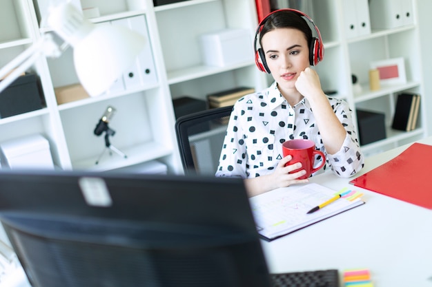 Una niña sentada en los auriculares en una mesa en la oficina, sostiene una taza roja en sus manos y mira el monitor.