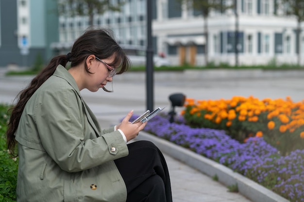 Una niña sentada en un banco en la calle con documentos en las manos y usando su teléfono.