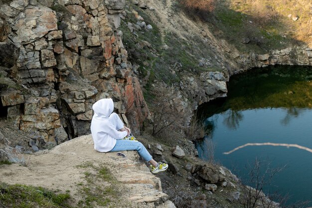 Niña sentada en el borde de una montaña con una sudadera con capucha blanca y mira el lago