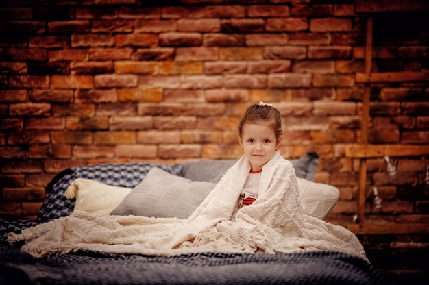 Una niña sentada en una cama gris envuelta en un plaid blanco frente a una pared de ladrillo marrón y mirando a puerta cerrada. Interior estilo loft. Copia espacio