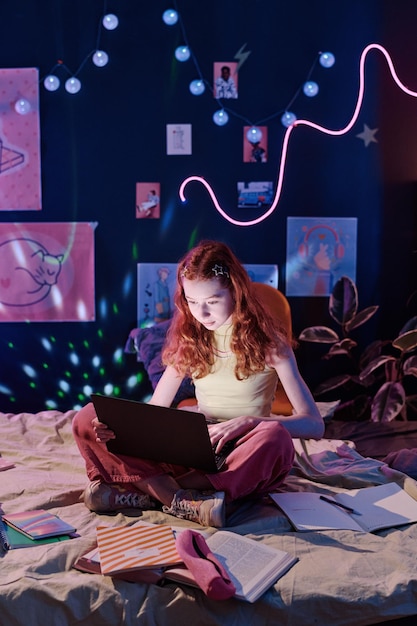 Foto niña sentada en la cama haciendo la tarea en la computadora portátil por la noche