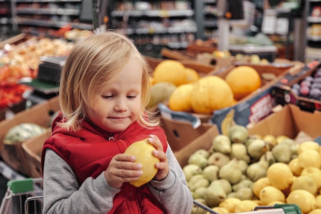 Niña sentada en un carrito de compras en una tienda de frutas o en un supermercado