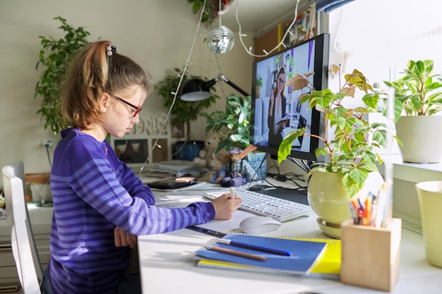 Niña sentada en casa en la mesa con una computadora escuchando una lección en línea