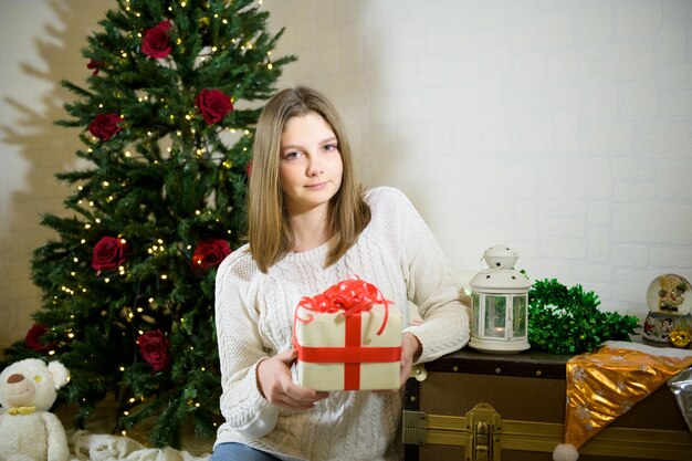 Niña sentada cerca de un abeto de Navidad y sostiene en sus manos un regalo