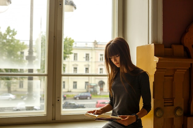 Foto niña sentada cerca de la ventana leyendo un libro