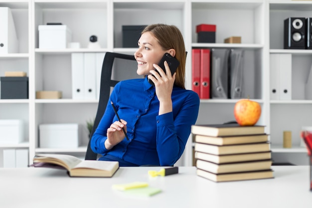 Una niña sentada en el escritorio de una computadora, con un lápiz en la mano y hablando por teléfono. Antes de que la niña mienta un libro abierto.